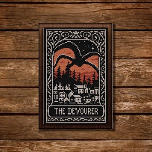 Pode incluir: Uma bordado em ponto cruz emoldurado com uma silhueta preta de uma criatura sobre uma cidade e uma floresta, com um fundo de pôr do sol vermelho e laranja. O texto "THE DEVOURER" está na parte inferior. A moldura é marrom, em um fundo de madeira.