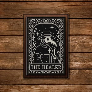 May include: A framed black and white cross-stitch depicting a plague doctor with a long beak, holding a staff and cross. The text "THE HEALER" is at the bottom. The artwork is set against a wooden background.