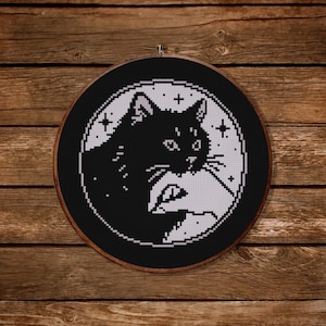 Puede incluir: Un diseño de punto de cruz en blanco y negro de un gato dentro de un círculo, sobre un fondo negro. El gato mira hacia la derecha, con una forma de media luna detrás. El diseño está enmarcado en un aro de madera.