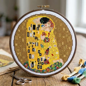 May include: A cross-stitch embroidery of Gustav Klimt's "The Kiss" in a wooden hoop. The artwork features a gold and yellow color palette with black, red, and floral accents. The embroidery is displayed on a wooden surface.
