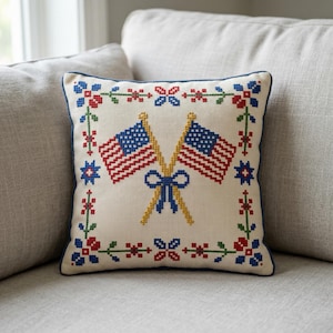 May include: Square decorative pillow with a cream-colored background. The pillow features a cross-stitch design of two American flags crossed with a yellow bow. The border is decorated with red, blue, and green floral patterns. The pillow is on a gray sofa.