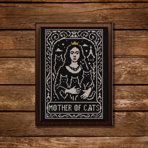 Può includere: Arte a punto croce in bianco e nero in una cornice di legno scuro. L'opera presenta una donna incoronata che tiene tre gatti, con la scritta "MOTHER OF CATS" in basso. Lo sfondo è nero con dettagli bianchi.