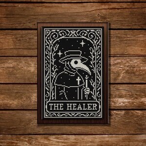 May include: A framed black and white cross-stitch depicting a plague doctor with a long beak, holding a staff and cross. The text "THE HEALER" is at the bottom. The artwork is set against a wooden background.