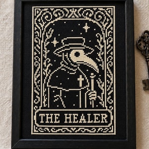 Puede incluir: Un bordado de punto de cruz en negro y crema dentro de un marco negro que muestra a un médico de la peste con sombrero de ala ancha y máscara de pico largo. El diseño incluye estrellas, una cruz y el texto THE HEALER en un borde decorativo.