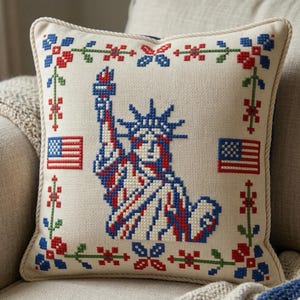 Peut inclure: Coussin carré décoratif avec un motif au point de croix de la Statue de la Liberté en rouge, blanc et bleu. Le coussin a un fond crème avec une bordure florale et deux drapeaux américains.