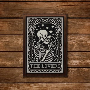 May include: Framed black and white cross-stitch art featuring two skeletons embracing, with hearts above them. The text "THE LOVERS" is at the bottom. The artwork is in a dark wooden frame, set against a wooden background.