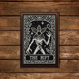 May include: A framed black and white cross-stitch featuring a Demogorgon-like creature with the text "THE RIFT". The artwork has an ornate border and is set against a wooden background. The design is detailed and evokes a vintage aesthetic.