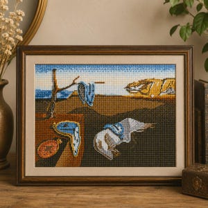 May include: Framed cross-stitch art depicting a surrealist landscape with melting clocks, inspired by Salvador Dalí. The artwork features a brown wooden frame, a beige mat, and a detailed stitched image with blue, brown, and beige tones.