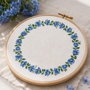 Peut inclure: Un tambour à broder circulaire en bois contenant un tissu blanc avec un motif au point de croix représentant une couronne de fleurs bleues aux centres jaunes et aux feuilles vertes. Le motif est disposé en cercle sur le tissu.