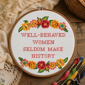 Puede incluir: Un bastidor de bordado de punto de cruz con la cita "WELL-BEHAVED WOMEN SELDOM MAKE HISTORY" en letras rojas. El diseño presenta un borde floral con flores amarillas, rojas y azules. El bastidor de madera es visible, y hilos de bordar y tijeras están en el fondo.