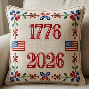 May include: Square decorative pillow with a cream-colored background. The pillow features the numbers "1776" and "2026" in red cross-stitch, along with two small American flags and floral embroidery in red, blue, and green.