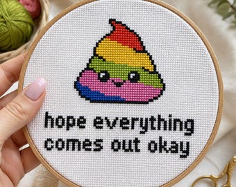 May include: Cross-stitch embroidery in a wooden hoop features a rainbow-colored poop emoji with a smiling face. The text "hope everything comes out okay" is stitched below the emoji. The hoop is held by a person.