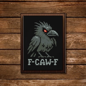 May include: Framed cross-stitch art featuring a pixelated raven with red eyes on a black background. The raven is gray and has a stern expression. The text "F-CAW-F" is stitched below the raven. The frame is dark brown, set against a wooden background.