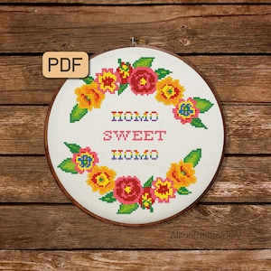 Könnte beinhalten: Ein weißer Stickrahmen mit einem floralen Kranzdesign in Gelb-, Rosa- und Grüntönen. Der Text "HOMO SWEET HOMO" ist in Regenbogenfarben in der Mitte des Rahmens gestickt.