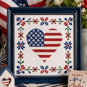 May include: A framed cross-stitch embroidery featuring a heart-shaped American flag pattern centered on white fabric, surrounded by a decorative border of red, blue, and green floral motifs, all encased in a dark blue wooden frame with gold trim.