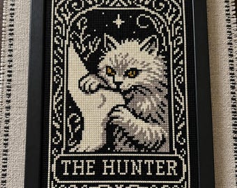 May include: A framed cross-stitch embroidery featuring a fluffy cat with yellow eyes clutching a white bird. The design is rendered in black, white, and gray threads against a black background, enclosed in an ornate border with the text THE HUNTER.