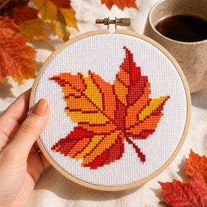 May include: A circular wooden embroidery hoop containing white fabric with a cross-stitch design of a single autumn leaf. The leaf features a gradient of vibrant colours including bright yellow, orange, and deep red, centred on the fabric.