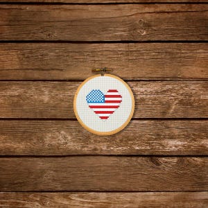 May include: A wooden embroidery hoop with a white fabric inside. The fabric has a red, white, and blue heart-shaped American flag design.