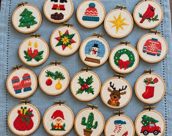 May include: A collection of twenty small circular wooden embroidery hoops, each featuring a unique cross-stitch design related to Christmas, including a tree, snowman, reindeer, cardinal, holly, ornaments, and a red truck carrying a tree.