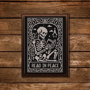 May include: Framed cross-stitch artwork in black and white, depicting a skeleton reading a book. The design includes a church and the text "READ IN PEACE". The frame is dark brown.