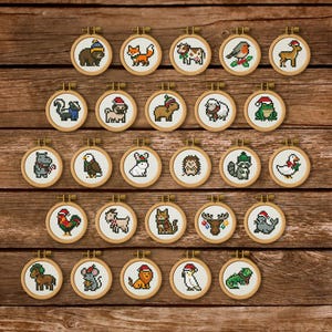 Puede incluir: Una colección de veinticinco pequeños bastidores de bordado circulares de madera, cada uno con un diseño único de punto de cruz en pixel art de un animal, incluyendo un oso, zorro, vaca, pájaro, ciervo, mofeta, pug, capibara, oveja, rana, hipopótamo, águila, conejo, erizo, mapache, pato, gallo, cabra, gato, alce, foca, caballo, ratón, león, loro y un lagarto.