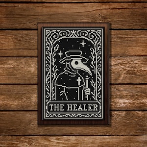 Könnte beinhalten: Ein gerahmtes Schwarz-Weiß-Kreuzstichbild, das einen Pestdoktor mit langem Schnabel zeigt, der einen Stab und ein Kreuz hält. Der Text "THE HEALER" steht unten. Das Kunstwerk befindet sich vor einem Holzgrund.
