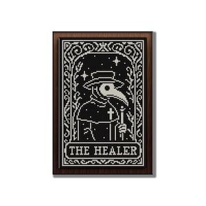 May include: A framed, black and white cross-stitch design featuring a plague doctor with a long beak, hat, and cross. The text "THE HEALER" is at the bottom. The frame is a dark brown color.