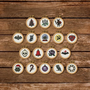 May include: A collection of nineteen circular wooden embroidery hoops, each containing a unique pixel art cross-stitch design. The motifs include a Christmas tree, candelabra, devil, bat, stocking, snow globe, deer, heart, skull, bell, rose, raven, and wreath.
