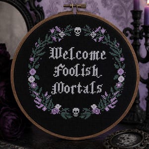 May include: A circular wooden embroidery hoop containing black fabric with a cross-stitch design. The text "Welcome Foolish Mortals" is stitched in white, surrounded by a floral wreath in shades of purple and green, with two small white skulls.