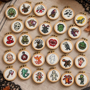Puede incluir: Una colección de pequeños bastidores de bordado circulares de madera con varios diseños de punto de cruz en pixel art. Los motivos incluyen monstruos, calaveras, fantasmas, animales y criaturas fantásticas dispuestos en filas sobre tela.