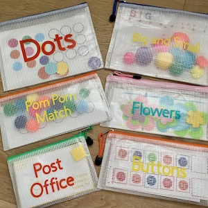 May include: Six rectangular clear mesh zipper pouches containing educational activity materials. The pouches are labeled with the text Dots, Big and Small, Pom Pom Match, Flowers, Post Office, and Buttons, each featuring colorful contents inside.