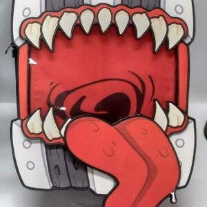 Puede incluir: Una mochila novedosa con forma de boca abierta de un monstruo de dibujos animados. El dise&ntilde;o presenta un interior rojo, dientes blancos y una larga lengua roja. El exterior tiene una combinaci&oacute;n de colores gris y blanco.