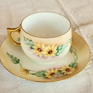 May include: A vintage teacup and saucer set. The cup and saucer are painted with yellow sunflowers and green leaves. The cup has a gold handle and trim. The saucer has a matching floral design and gold trim. The set is on a cream-colored surface.
