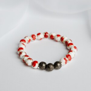 May include: A beaded bracelet featuring white and red floral patterned beads interspersed with three dark gray, textured beads. The bracelet is displayed on a white background.