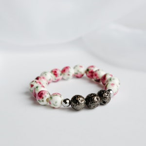 May include: A bracelet with white beads decorated with a pink floral design, combined with dark grey lava beads and a silver bead. The bracelet is displayed on a white surface.