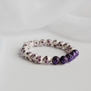 May include: A bracelet with white beads adorned with purple floral designs, transitioning to deep purple beads. The jewellery is displayed against a white backdrop, highlighting the colour contrast. A fashionable accessory.