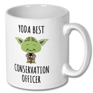 May include: White ceramic mug featuring a green cartoon Yoda character holding a stick. The text "YODA BEST CONSERVATION OFFICER" is printed on the mug.