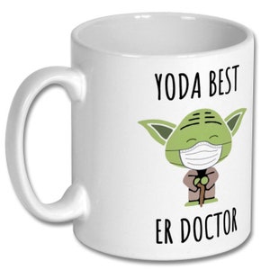 Może przedstawiać: Biały kubek ceramiczny z zieloną postacią z kresk&oacute;wki, kt&oacute;ra nosi maskę na twarz i trzyma laskę. Na kubku nadrukowany jest tekst "YODA BEST ER DOCTOR".