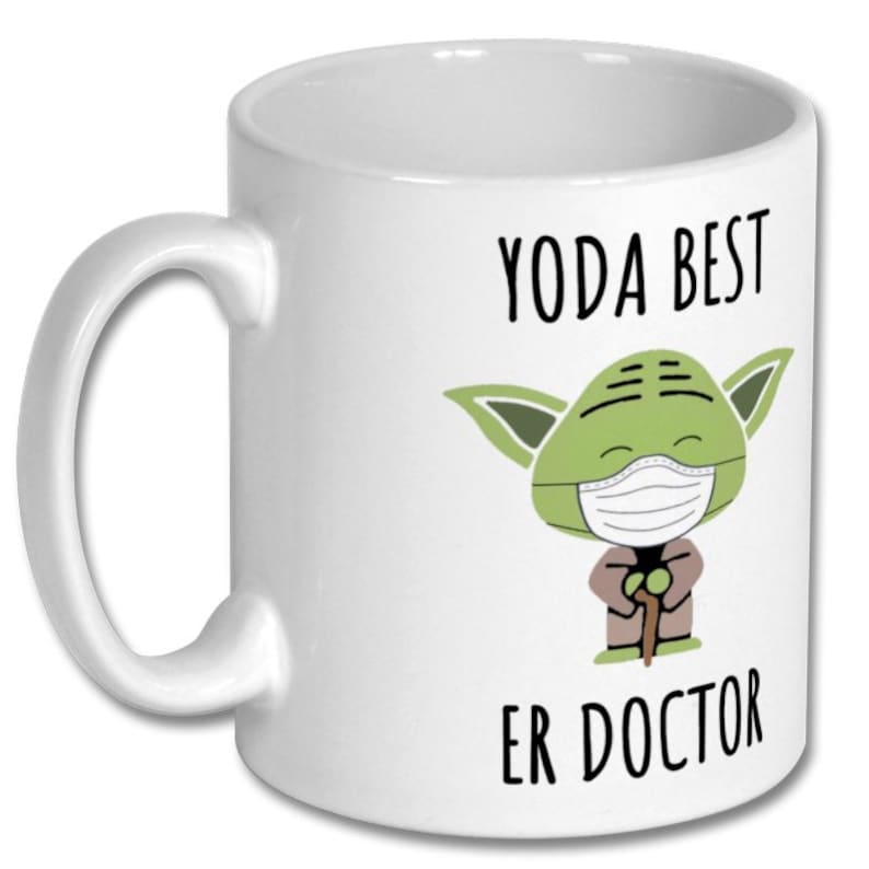 Może przedstawiać: Biały kubek ceramiczny z zieloną postacią z kresk&oacute;wki, kt&oacute;ra nosi maskę na twarz i trzyma laskę. Na kubku nadrukowany jest tekst "YODA BEST ER DOCTOR".
