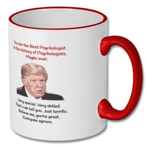 May include: White ceramic coffee mug with a red rim and handle. The mug features a cartoon image of Donald Trump and text that reads "You are the Best Psychologist in the history of Psychologists. Maybe ever. Very special. Very skilled. That I can tell you. Just terrific. Believe me, you're great. Everyone agrees."