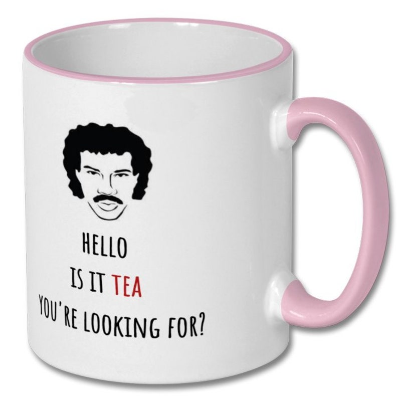 May include: White ceramic mug with a pink rim and handle. The mug features a black and white illustration of a man with a moustache and the text "HELLO IS IT TEA YOU'RE LOOKING FOR?"