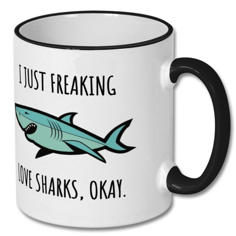 Puede incluir: Taza de caf&eacute; de cer&aacute;mica blanca con borde y asa negros. La taza tiene un tibur&oacute;n de dibujos animados y el texto "Me encantan los tiburones, &iquest;vale?"