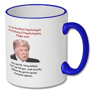 May include: White ceramic coffee mug with a blue rim and handle. The mug features a cartoon image of Donald Trump and text that reads "You are the Best Psychologist in the history of Psychologists. Maybe ever. Very special. Very skilled. That I can tell you. Just terrific. Believe me, you're great. Everyone agrees."
