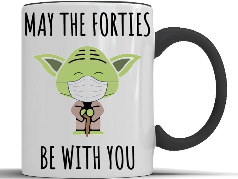 May include: White ceramic mug with a black handle. The mug features a green cartoon character wearing a face mask and holding a walking stick. The text "May the Forties Be With You" is printed on the mug.
