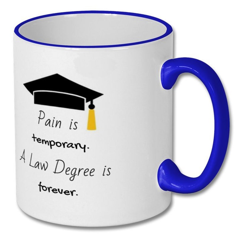May include: White ceramic mug with a blue handle and rim. The mug features a black graduation cap graphic with a yellow tassel and the text "Pain is temporary. A Law Degree is forever."