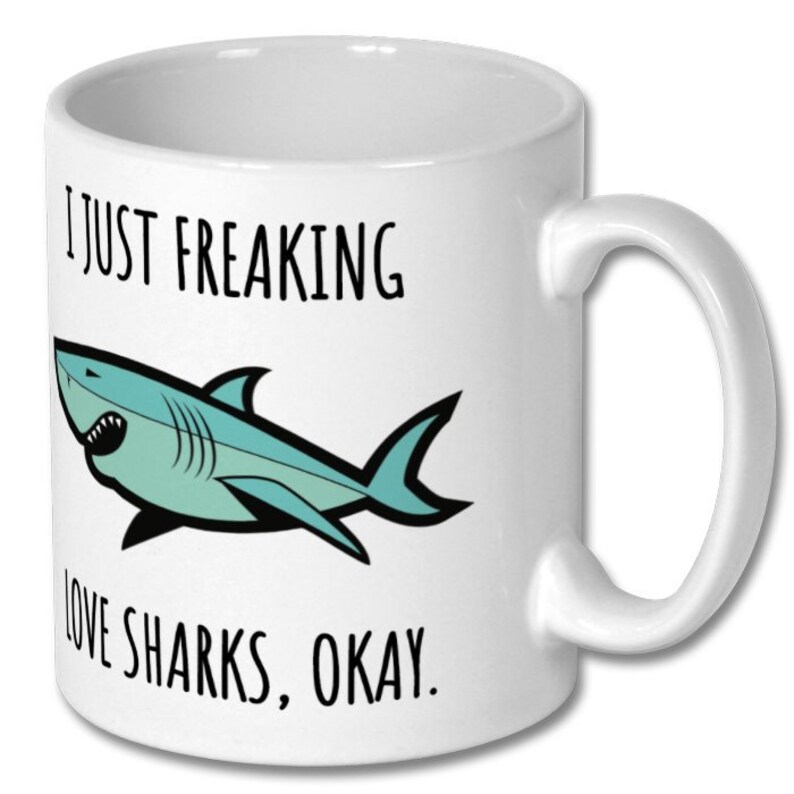 Puede incluir: Taza de caf&eacute; de cer&aacute;mica blanca con un tibur&oacute;n azul de dibujos animados y el texto "Me encantan los tiburones, &iquest;vale?"
