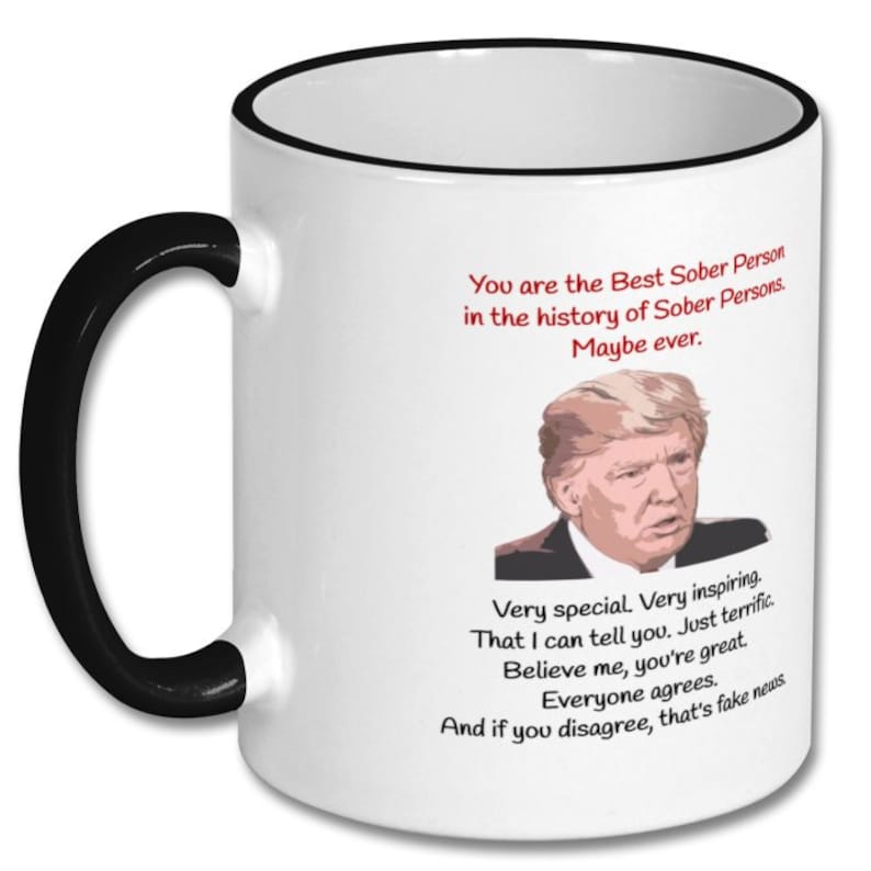 May include: White coffee mug with a black rim and handle. The mug features a humorous image of a man with the text "You are the Best Sober Person in the history of Sober Persons. Maybe ever. Very special. Very inspiring. That I can tell you. Just terrific. Believe me, you're great. Everyone agrees. And if you disagree, that's fake news."