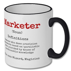 May include: White ceramic coffee mug with a black rim and handle. The mug has a red and black design with the word "Marketer" and a definition of the word. The definition reads: "Someone who does precision work based on unreliable data provided by those of questionable knowledge. Also see: Wizard, Magician."