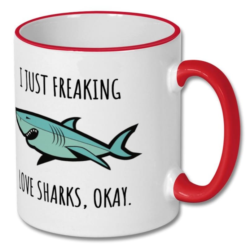 Puede incluir: Taza de caf&eacute; de cer&aacute;mica blanca con borde y asa rojos. La taza presenta un tibur&oacute;n de dibujos animados y el texto "Me encantan los tiburones, &iquest;vale?"