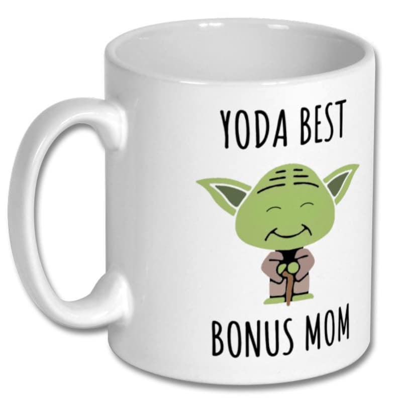 K&ouml;nnte beinhalten: Wei&szlig;er Keramikbecher mit Henkel. Der Becher zeigt ein Cartoon-Bild von Yoda mit dem Text "YODA BEST BONUS MOM" in Schwarz. Die Yoda-Figur ist gr&uuml;n mit einem braunen Gewand und einem Gehstock.